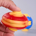 A close-up of a hand holding a red and yellow plastic stress ball toy, with a second blue and yellow toy blurred in the background against a light gray backdrop. Fidgit Sphere Spinner