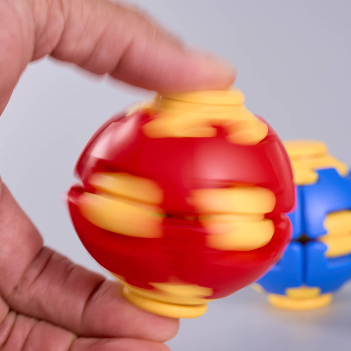 A close-up of a hand spinning a red and yellow plastic ball toy, with a second blue and yellow ball blurred in the background against a light gray, Fidgit Sphere Spinner backdrop.