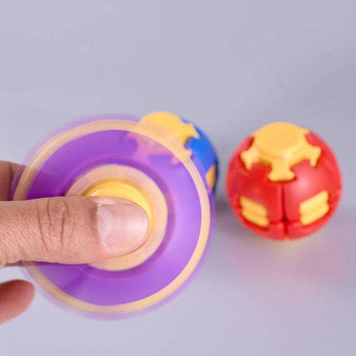 A close-up of a hand spinning a purple and yellow gyroscopic toy, with a red and yellow ball toy blurred in the background against a light gray surface. Fidgit Sphere Spinner