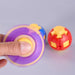 A close-up of a hand spinning a purple and yellow gyroscopic toy, with a red and yellow ball toy blurred in the background against a light gray surface. Fidgit Sphere Spinner