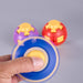 A hand spinning a blue and yellow plastic fidget spinner in the foreground, with two colorful toy eggs in the blurred background on a light gray surface. Fidgit Sphere Spinner