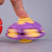 A purple and yellow plastic toy gyroscope spinner being spun by a human finger, shown against a neutral gray background. Fidgit Sphere Spinner