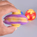 A hand holding a purple and yellow plastic spinning top toy, with two more toy tops blurred in the background on a light gray surface. Fidgit Sphere Spinner