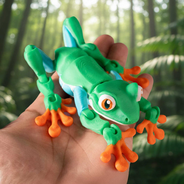 A green and blue tree frog toy with orange feet is held in a person's hand against a blurred green forest background.