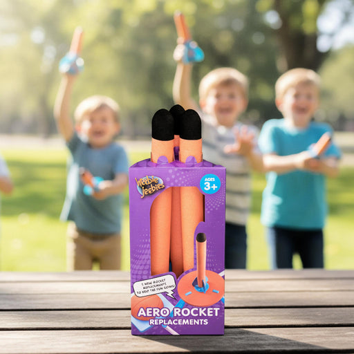 A purple retail box of Rocket Verbe Aerodynamic Rocket Replacements stands on a wooden table outdoors, with three blurred children holding orange rockets in a park behind it.