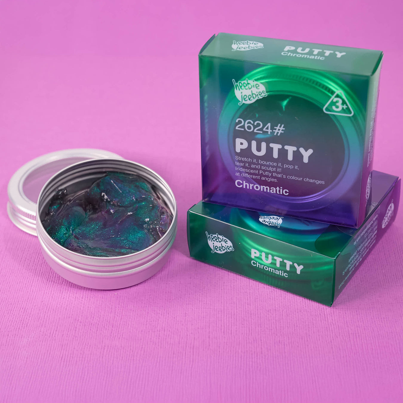 A round metal tin of Putty Chromatic 2624 putty, shown open with the lid beside it, next to two green and purple product boxes on a pink background.