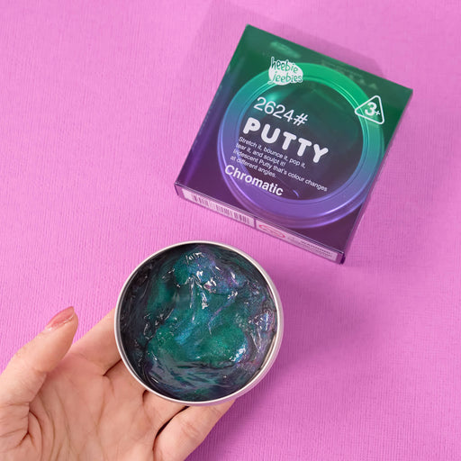A hand holds an open round tin of Chromatic 2624# Putty, showing the glossy dark teal and blue putty inside, with its purple and green packaging box on a solid pink background.
