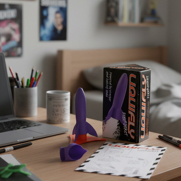 A Heebie Jeebies purple and orange rocket-shaped instructions displayed on a wooden desk next to its black and orange retail box, with a laptop, mug, and bedroom background visible.
