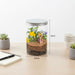 A clear glass jar terrarium with a silver metal lid sits on a light wood desk. The jar contains brown soil, green leaves, yellow and purple flowers, and a small fairy figurine.