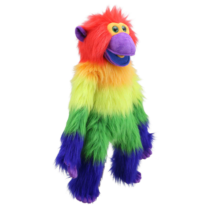 A plush rainbow monkey toy sits on a light wood table in a bright children's room. The toy has fluffy fur with red, orange, yellow, green, and purple color-blocked sections, large yellow eyes, and a purple snout and feet.