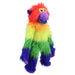 A plush rainbow monkey toy sits on a light wood table in a bright children's room. The toy has fluffy fur with red, orange, yellow, green, and purple color-blocked sections, large yellow eyes, and a purple snout and feet.
