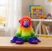 A plush rainbow monkey toy sitting on a light wood table in a softly blurred children's room. The toy has bright red hair, a purple face and ears, a yellow-green body, green legs, and purple feet and hands.