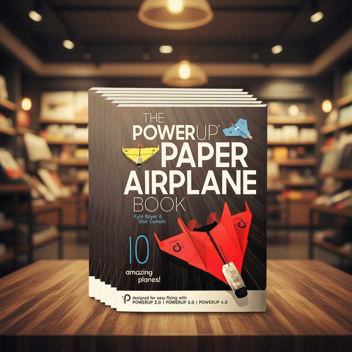 Stack of 'The PowerUp Paper Airplane Book' on a wooden table with bookshelves in the background
