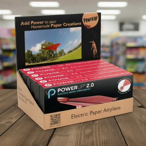 Display box of PowerUp 2.0 Electric Paper Airplane Kit on a wooden surface with a blurred store background.