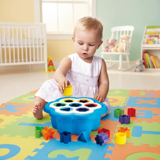 A toddler sits on a colorful foam play mat in a bright nursery, playing with a blue flower-shaped sorting toy and colorful geometric blocks.