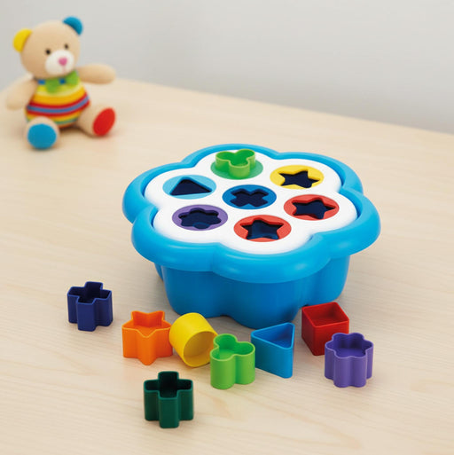 A colorful plastic shape-sorting toy set on a light wood surface, with a blue flower-shaped tray holding multicolored geometric pieces and additional loose pieces arranged around it. A small teddy bear toy sits blurred in the background.