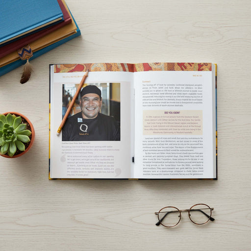 An open book or magazine laid flat on a light wood surface, showing printed text and a portrait photo of a smiling man. A pencil rests on the pages, with stacked books, a small succulent, and eyeglasses around it.