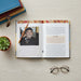 An open book or magazine laid flat on a light wood surface, showing printed text and a portrait photo of a smiling man. A pencil rests on the pages, with stacked books, a small succulent, and eyeglasses around it.
