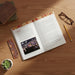 An open book laid on a wooden desk with printed text and a small embedded photo on the left page. The book has a decorative multicolor patterned cover and matching patterned interior accents.